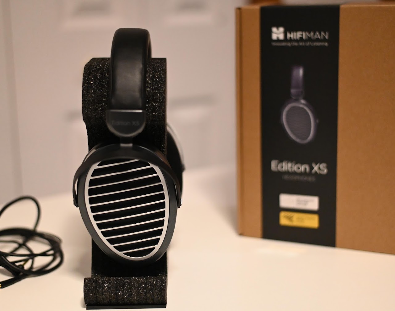 Front view of Hifiman Edition XS headphones on a foam stand beside their box